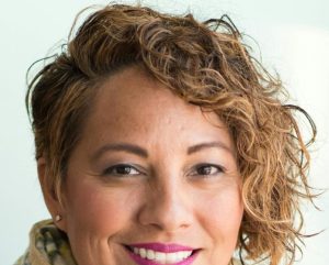 Smiling woman in a purple dress and scarf holding a tablet indoors.